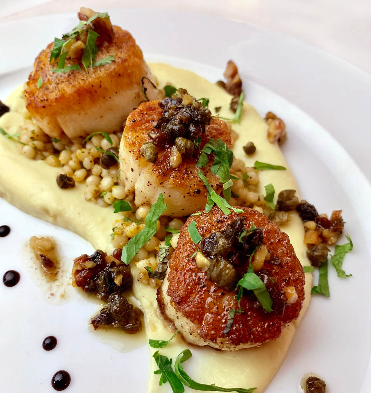 Three seared scallops garnished with herbs and capers, served on a bed of creamy sauce with small grains on a white plate.