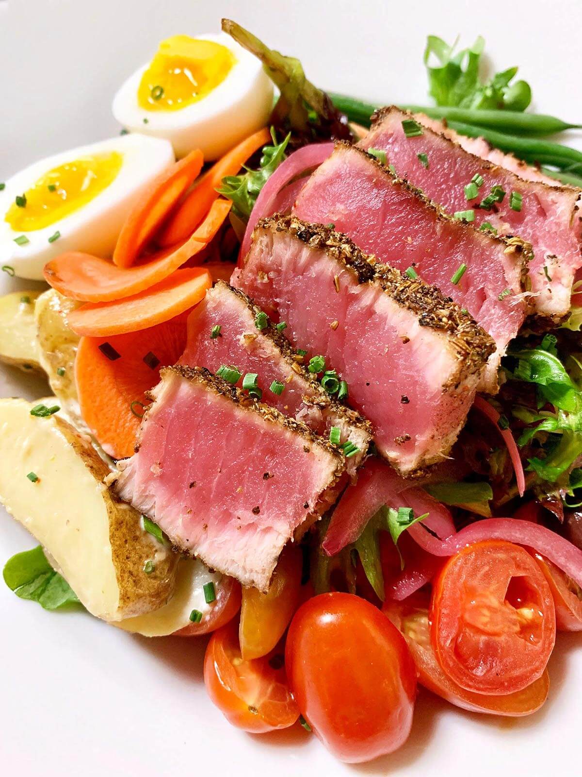 Seared rare tuna slices over mixed salad with cherry tomatoes, carrot slices, boiled potato wedges, and halved soft-boiled eggs.