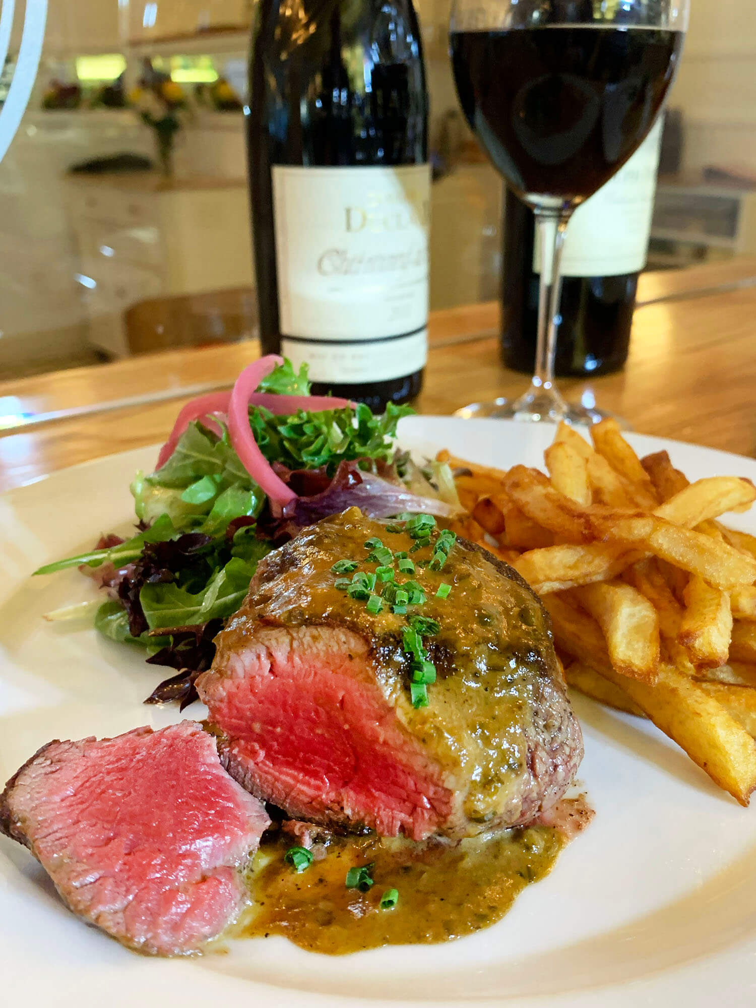Plate with medium-rare steak topped with sauce and chives, served with mixed green salad and crispy French fries, with wine bottles and glass in the background.