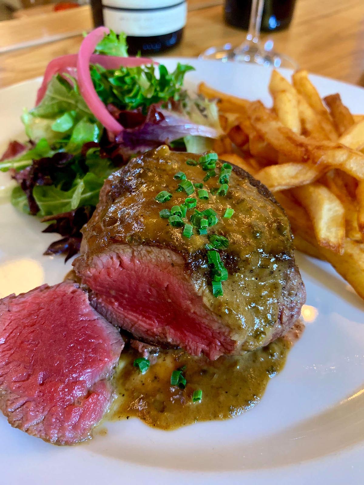 Plate with medium-rare steak topped with green herbs and sauce, side salad with greens and pickled onions, and French fries.