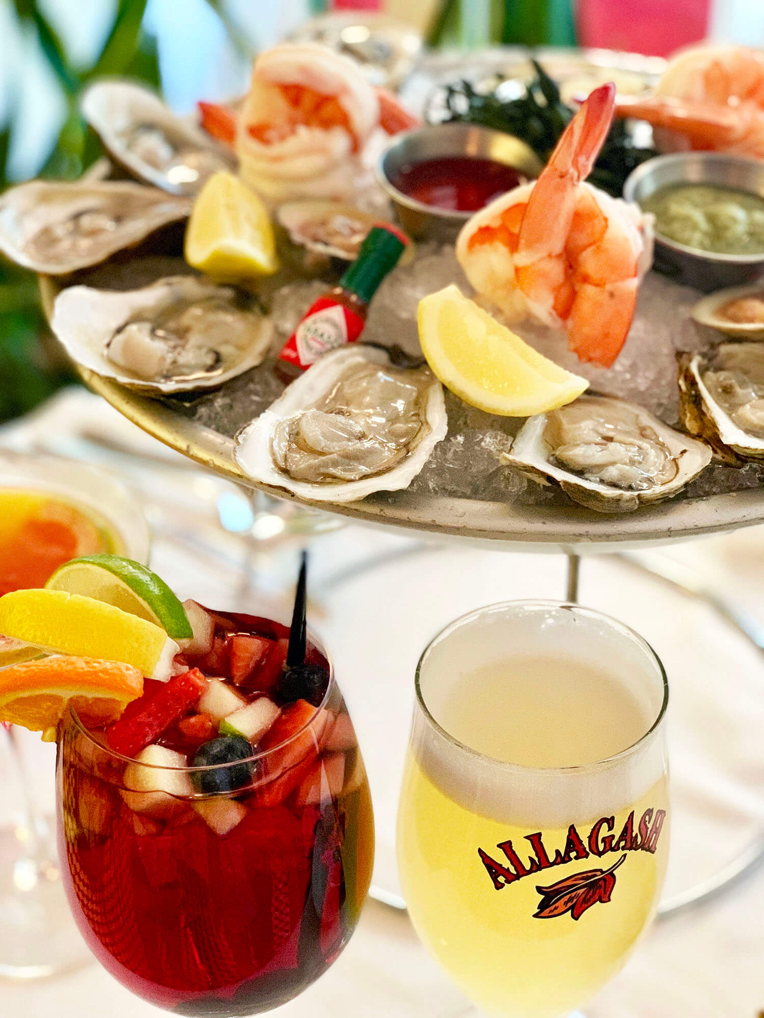 Plate of oysters on ice with lemon wedges, shrimp, cocktail sauce, and horseradish sauce, accompanied by a red sangria and a glass of Allagash beer.