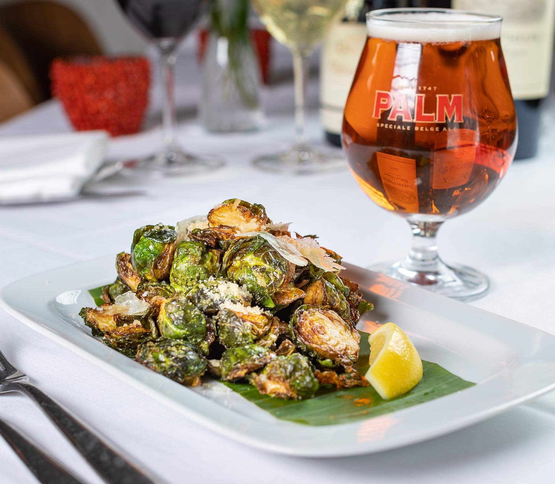 Crispy roasted Brussels sprouts garnished with shaved Parmesan and served with a lemon wedge on a white plate, next to a glass of amber beer.