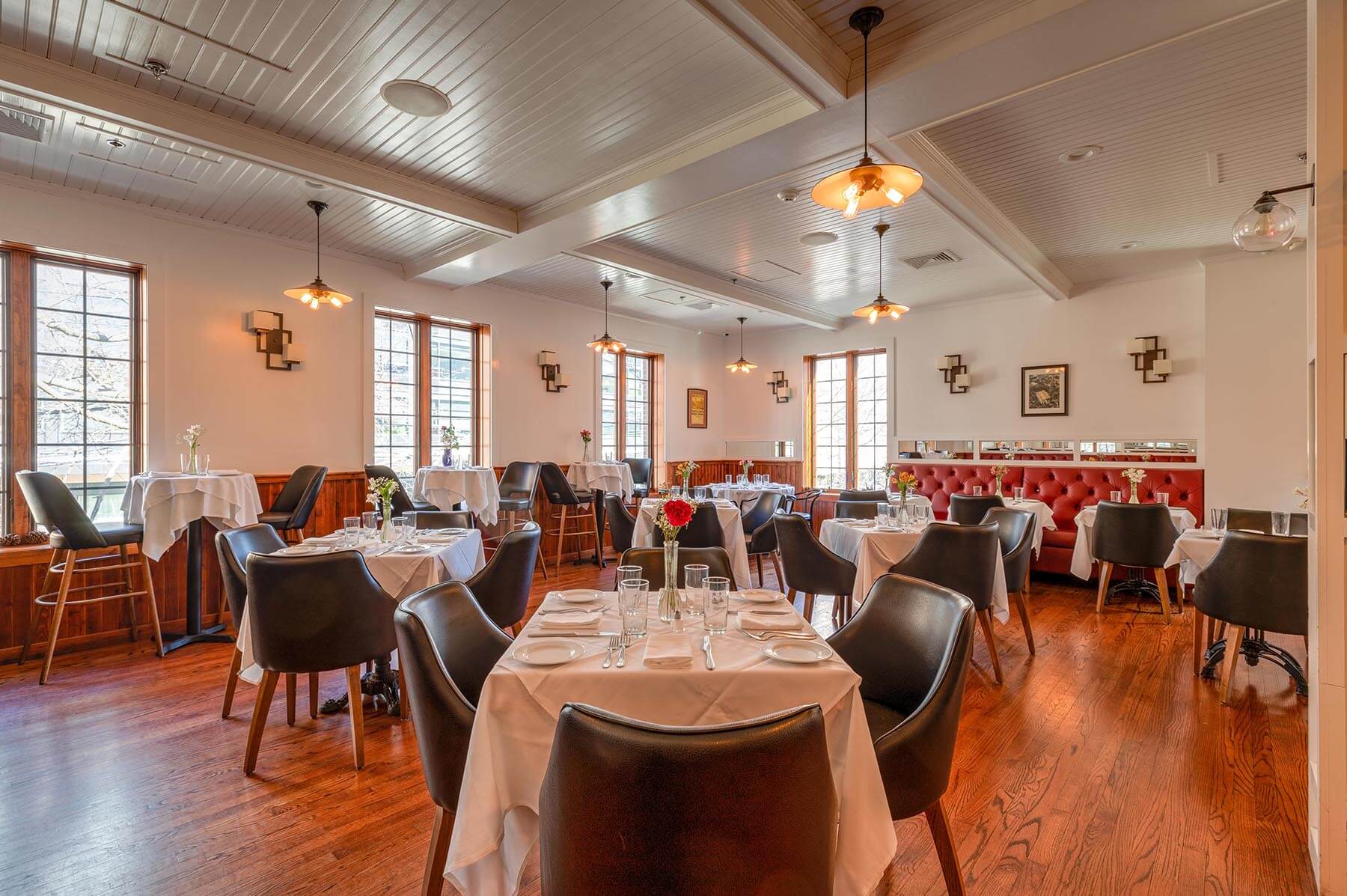 Well-lit elegant restaurant interior with wooden floor, white tablecloths, black chairs, and pendant lights.