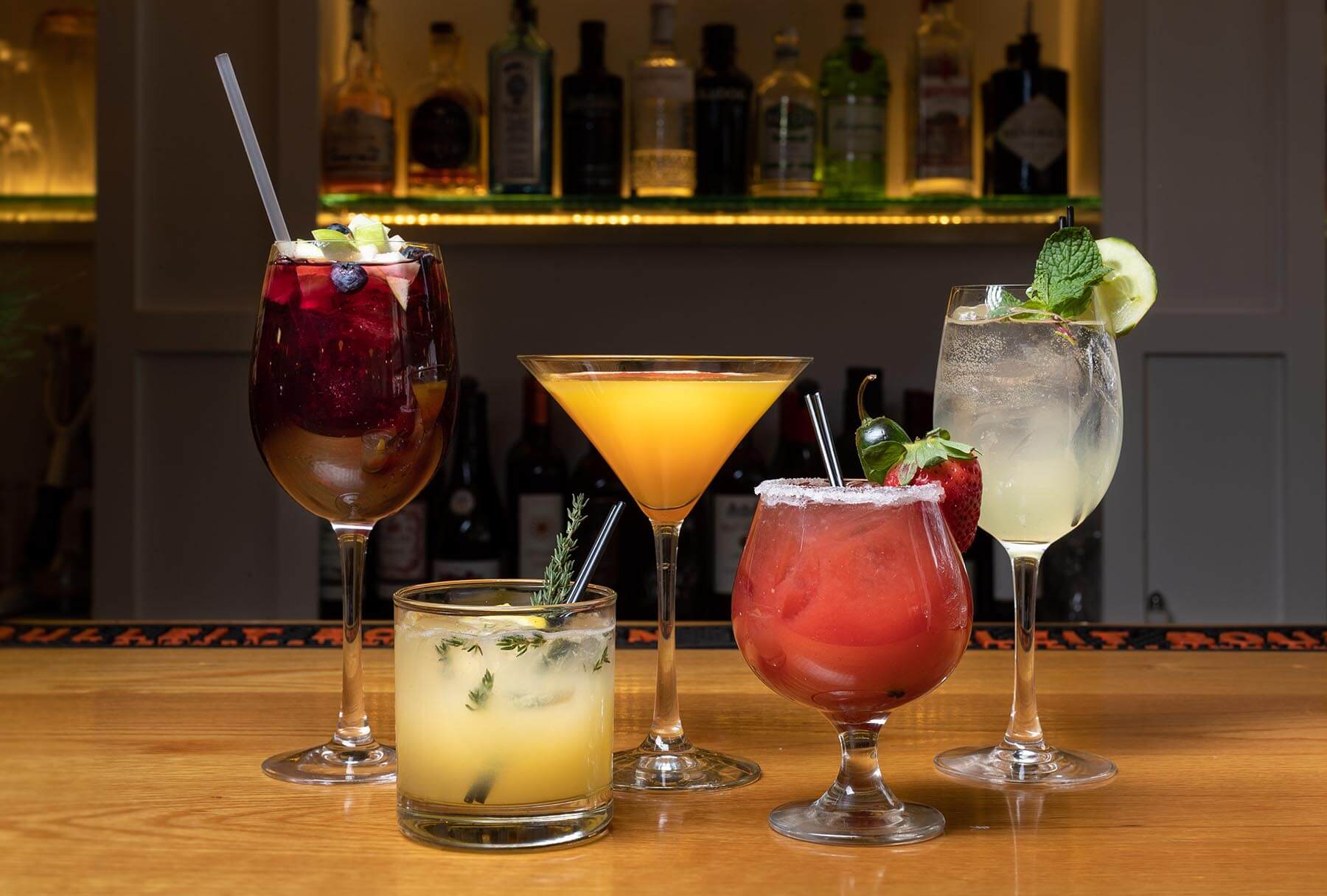 Five colorful cocktails on a wooden bar counter with garnishes and a background of liquor bottles.