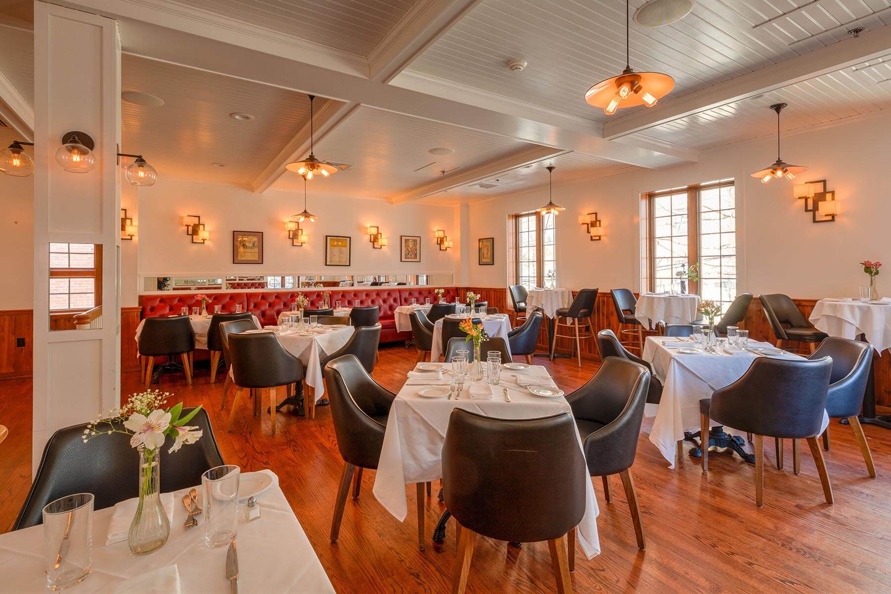 Cozy restaurant interior with wooden floors, white tablecloth-covered tables, black chairs, and warm lighting fixtures.