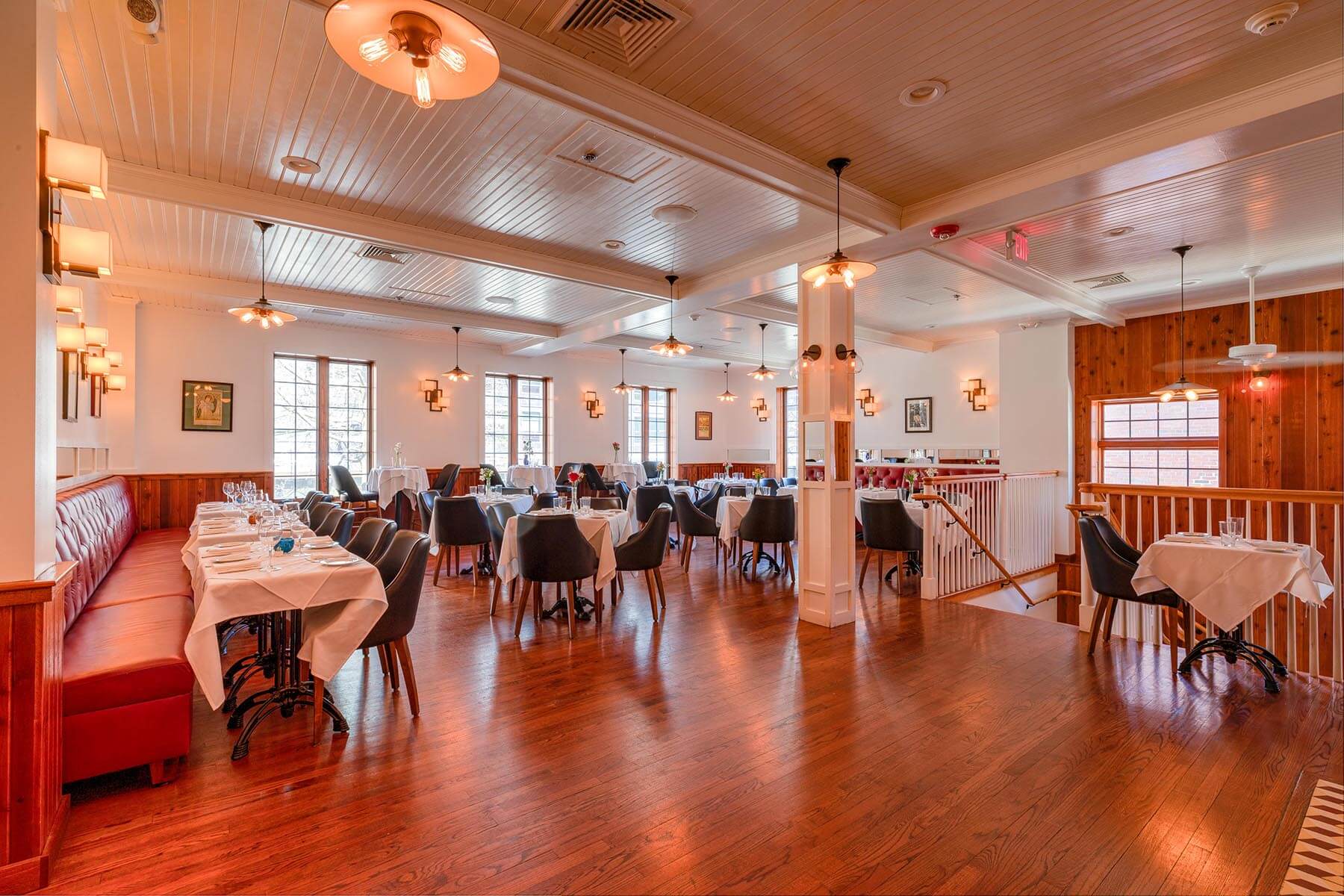 Bright restaurant interior with wooden floors, white walls, red banquette seating, and tables set with white linens and glassware.