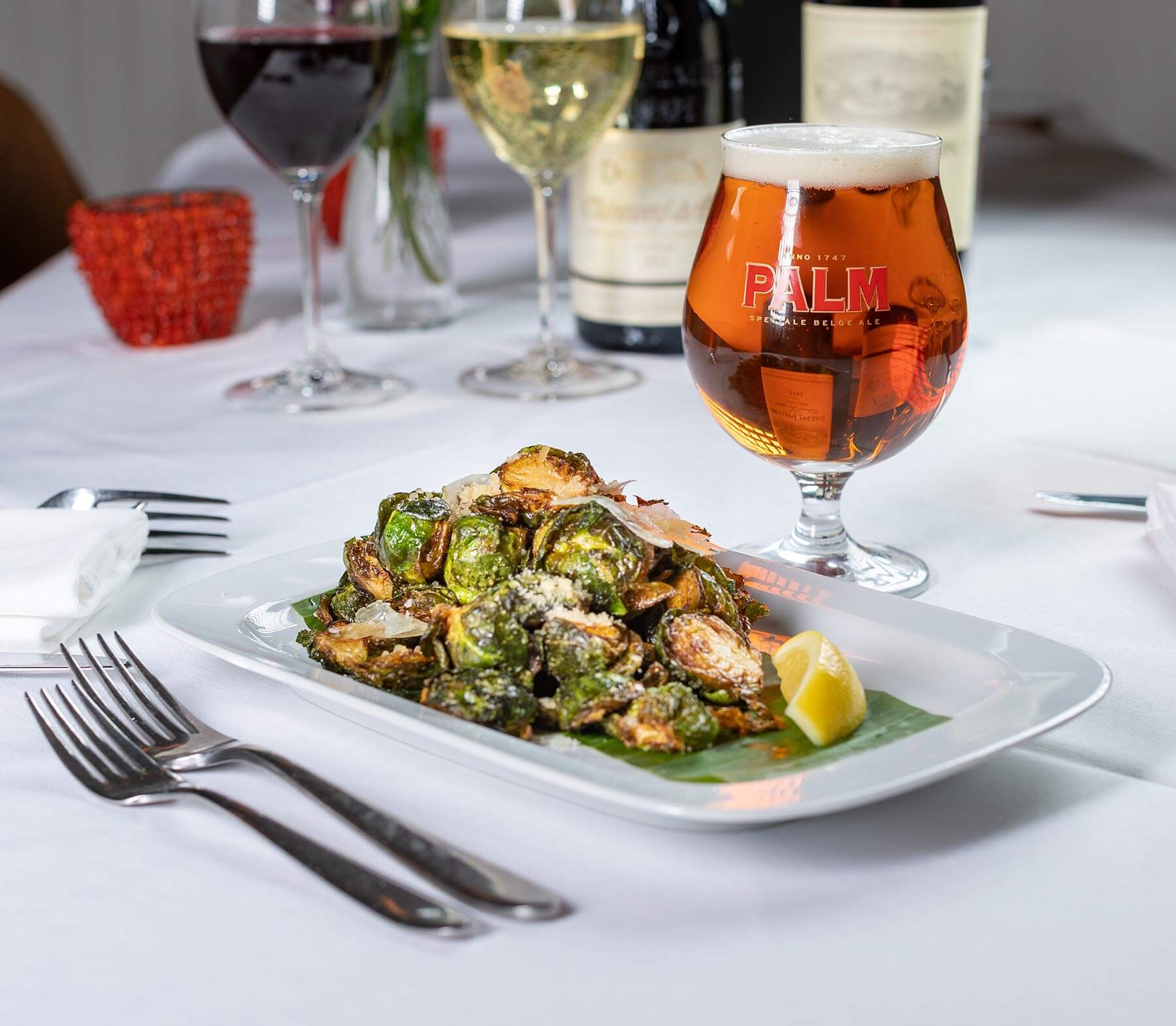 Plate of roasted Brussels sprouts with parmesan and lemon wedge beside a glass of amber Palm Belgian ale on a white tablecloth.