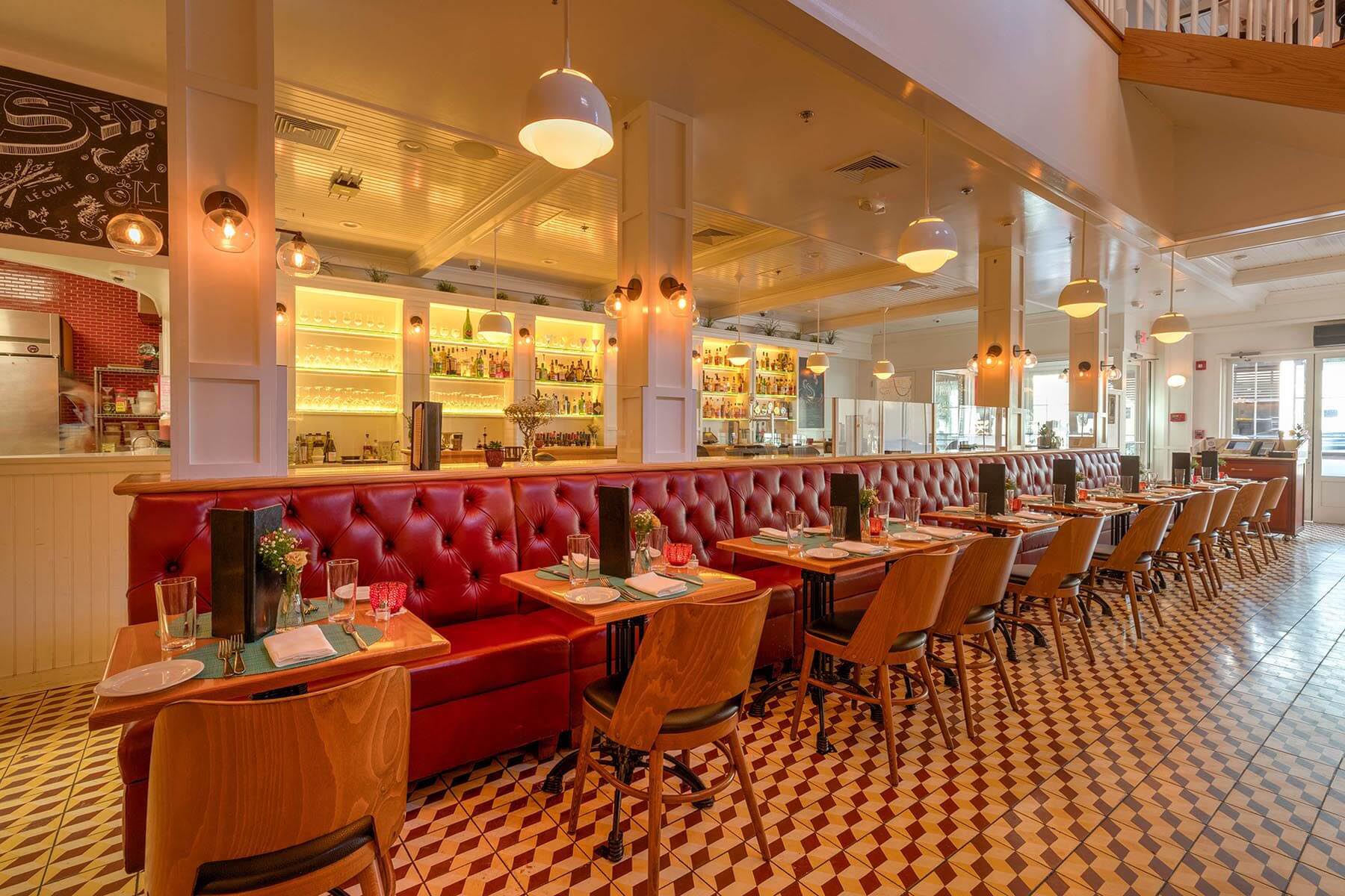 Bright restaurant interior with long red upholstered banquette seating, wooden tables set with plates and glasses, and warm pendant lighting.