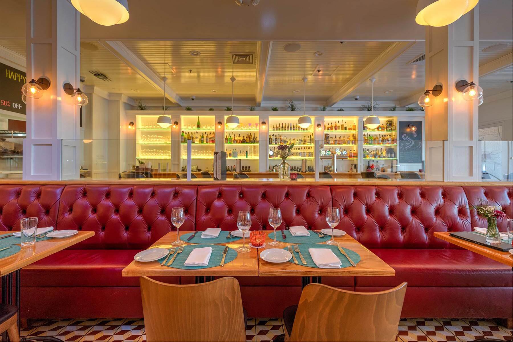 Elegant restaurant interior with red tufted banquette, wooden tables set with blue placemats, glassware, and white napkins, and a well-lit bar stocked with bottles in the background.
