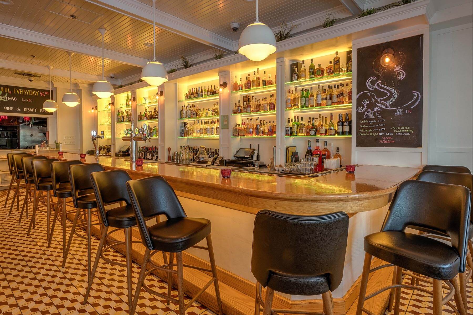 Interior of a stylish bar with a long wooden counter, black leather bar stools, and shelves of liquor bottles illuminated by warm lighting.