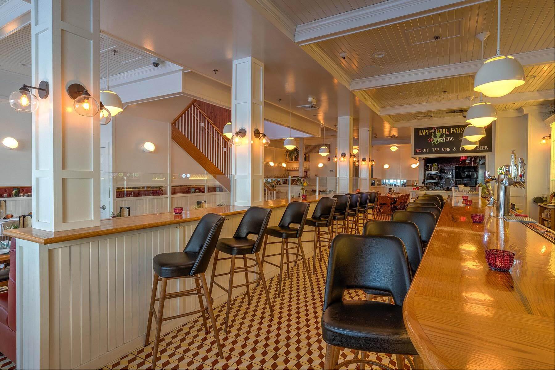 Interior of a well-lit bar with black cushioned bar stools lined up along a wooden bar counter and a checkered tile floor.