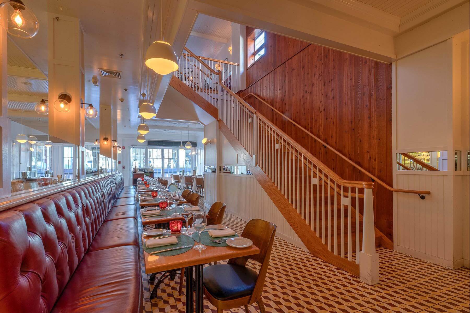 Interior of a restaurant with a long red leather bench, wooden tables set for dining, geometric tiled floor, and a wooden staircase with white railing.