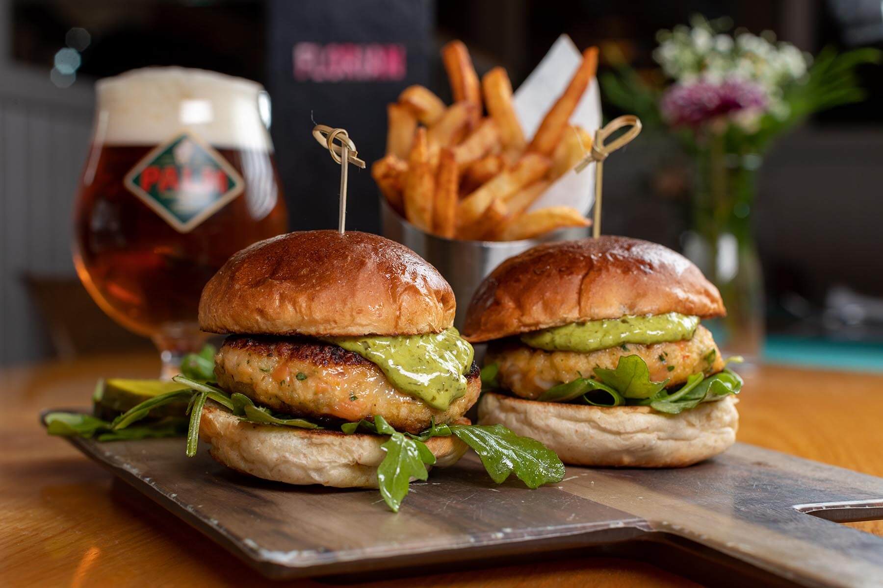 Two gourmet chicken burgers with green sauce and leafy greens on wooden serving board, frizzled French fries and glass of beer in background.