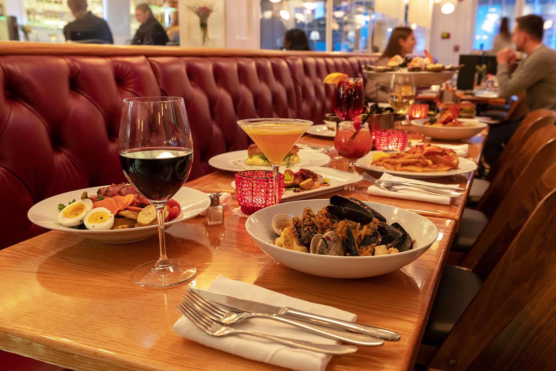 Table at a restaurant with plates of seafood, salad, fries, and various cocktails, including a glass of red wine and a martini glass, against a red leather bench.
