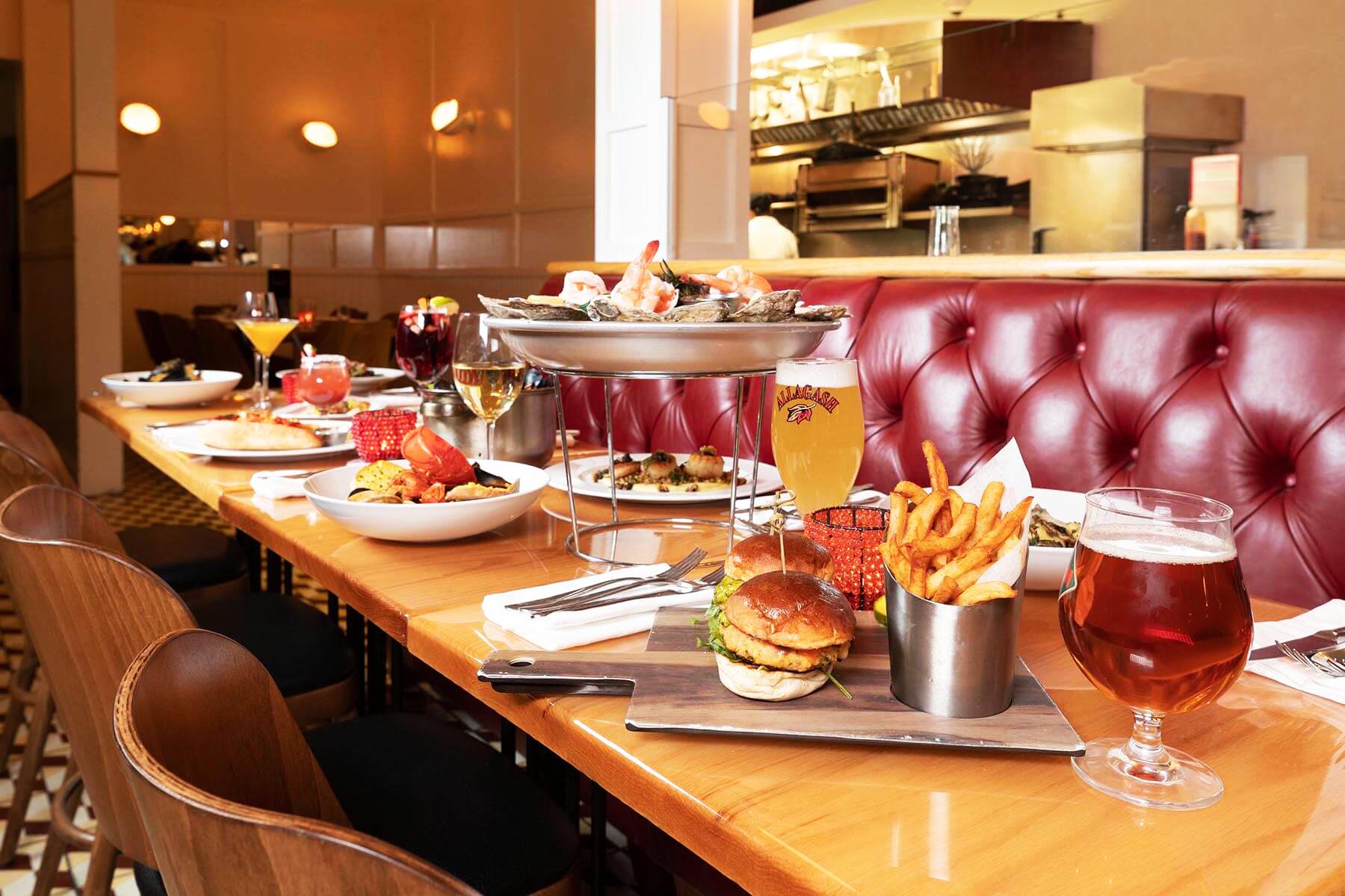 Restaurant table with seafood platter, sliders with fries, various drinks including beer and wine, and plates of food against a background of red leather booth seating.