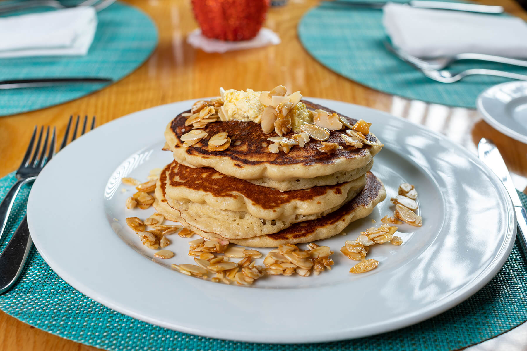 Stack of three golden brown pancakes topped with butter and almond slices on a white plate with a turquoise placemat.