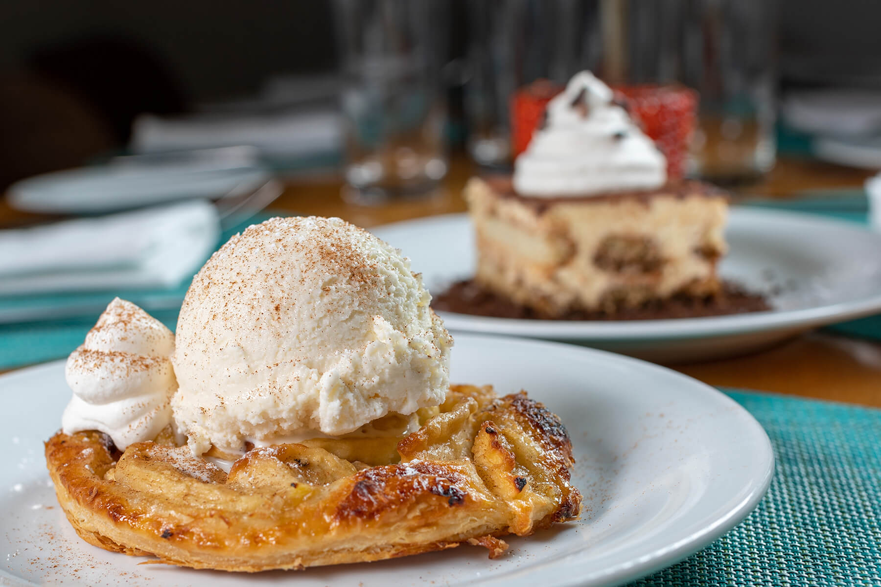 Close-up of a dessert plate featuring a warm banana pastry topped with a scoop of vanilla ice cream and a dollop of whipped cream, with another dessert in the background.