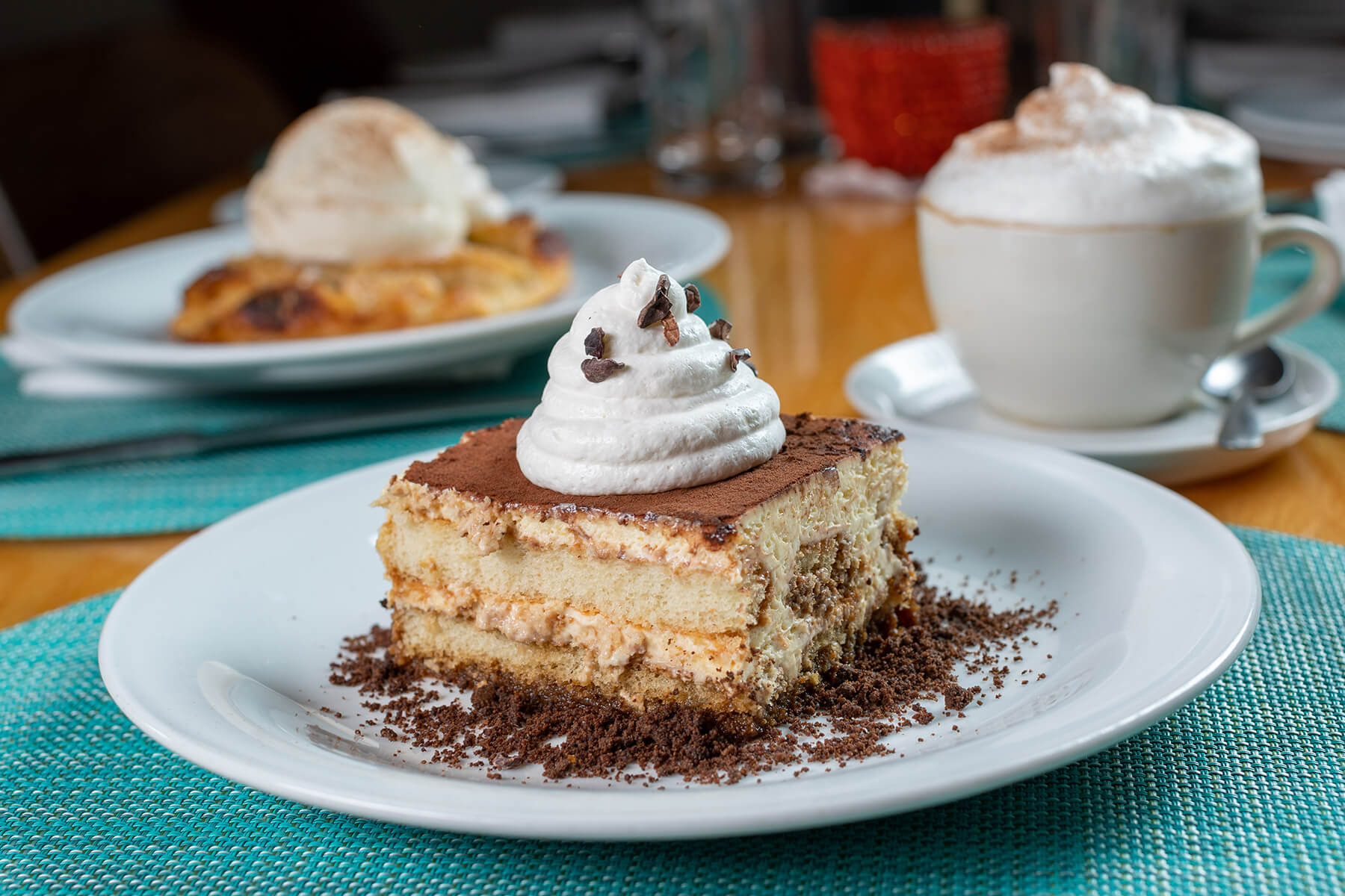 Slice of tiramisu topped with whipped cream and chocolate shavings on a white plate with a coffee cup and another dessert in the background.