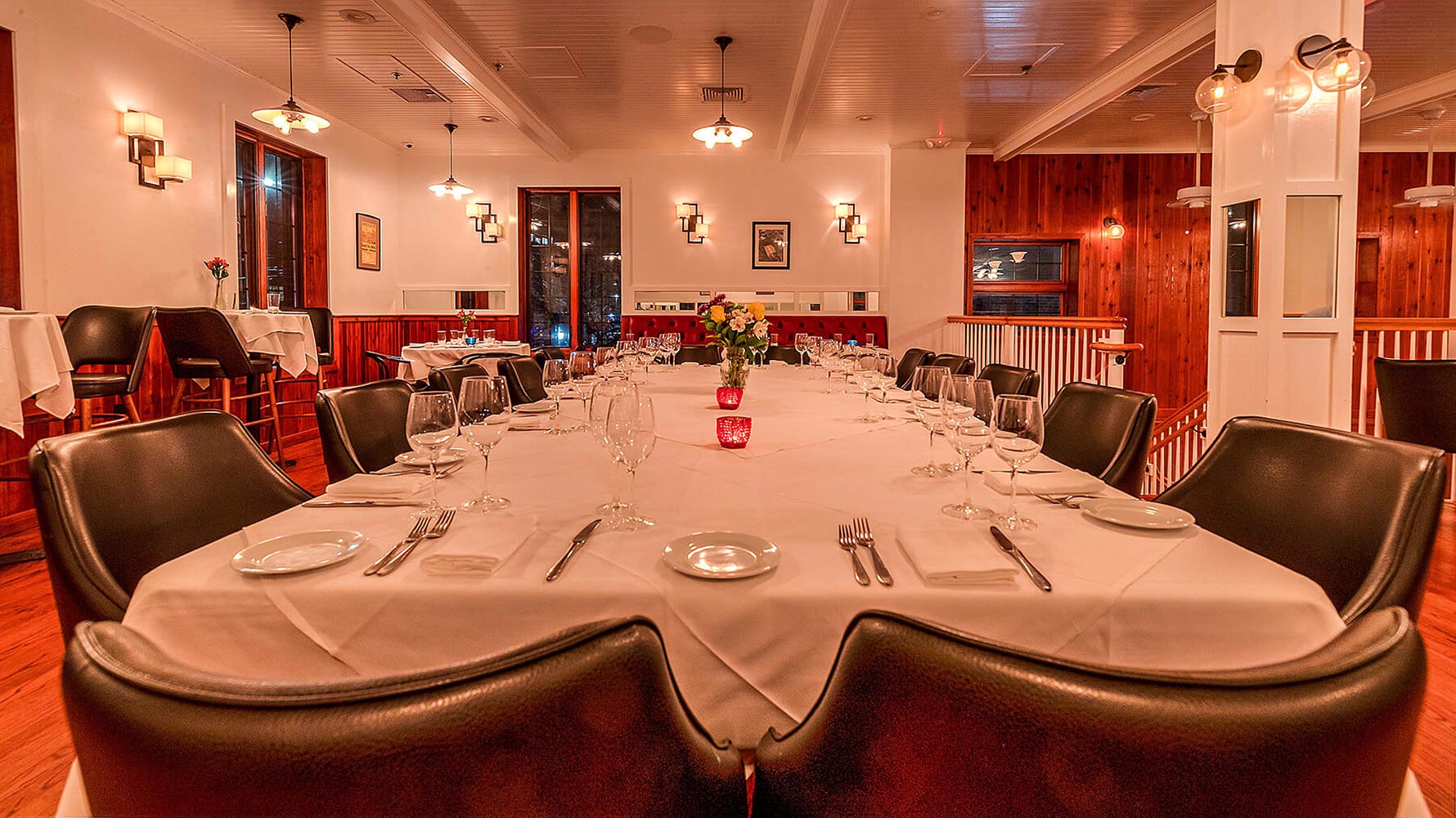 Elegant dining room with a long table set for a formal meal, featuring white tablecloths, dark leather chairs, glassware, and a floral centerpiece.