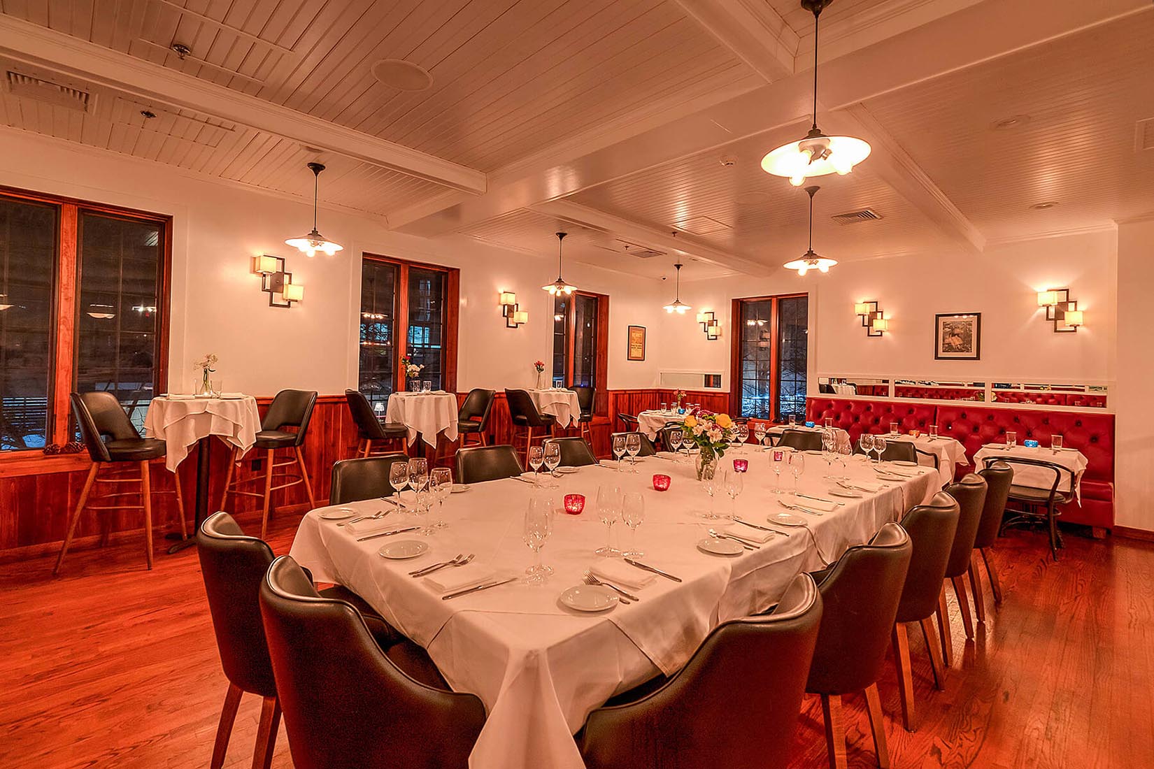 Warmly lit elegant dining room with a large rectangular table set for a meal, surrounded by leather chairs, smaller tables with white tablecloths, and red cushioned bench seating.