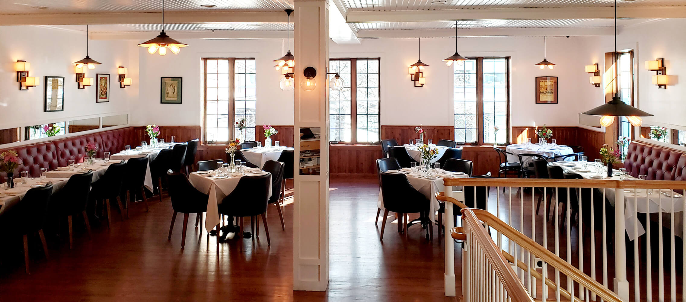 Bright and elegant restaurant dining area with round tables covered in white tablecloths, black chairs, red tufted bench seating, pendant lights, and large windows.