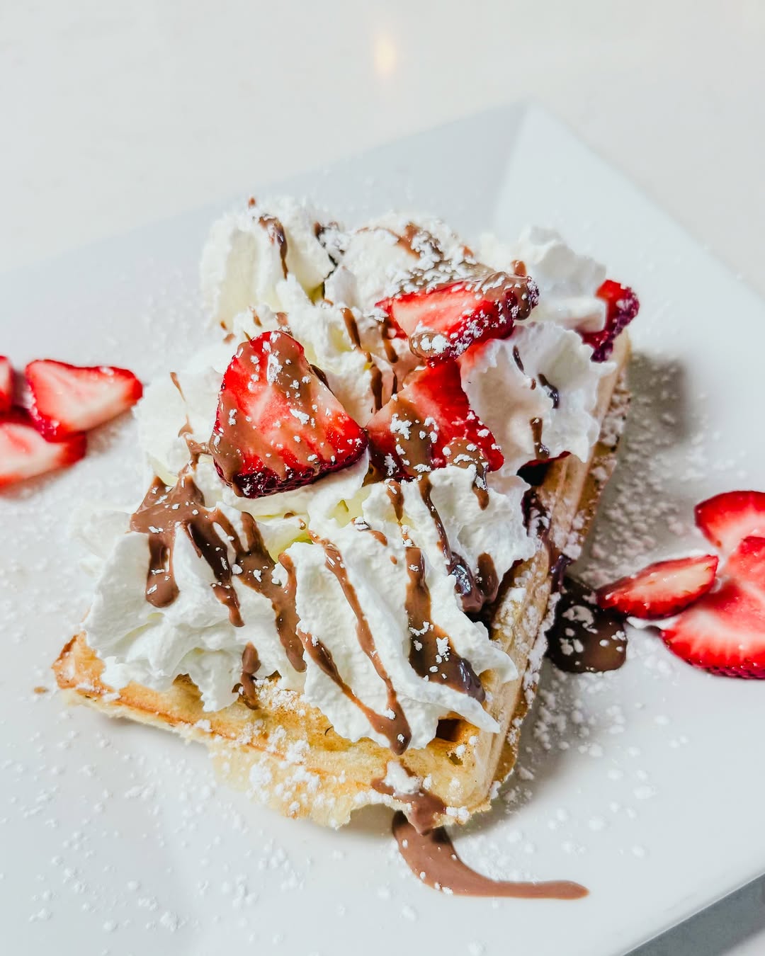 Waffle topped with whipped cream, sliced strawberries, chocolate drizzle, and powdered sugar on a white plate.