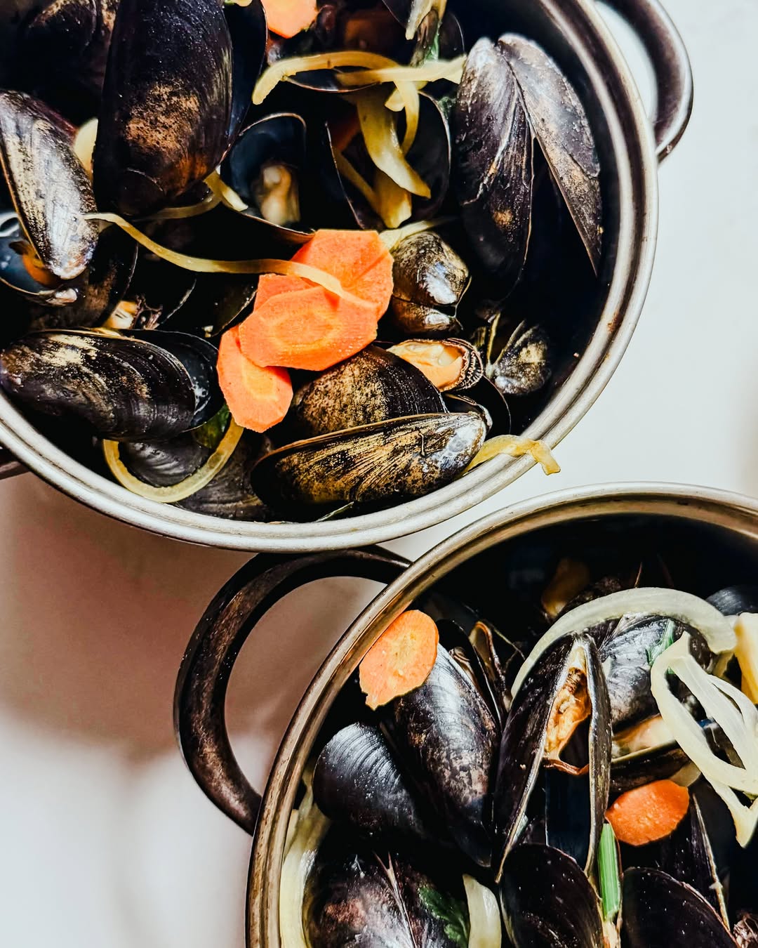 Two metal pots filled with cooked mussels, sliced carrots, and onions on a white surface.