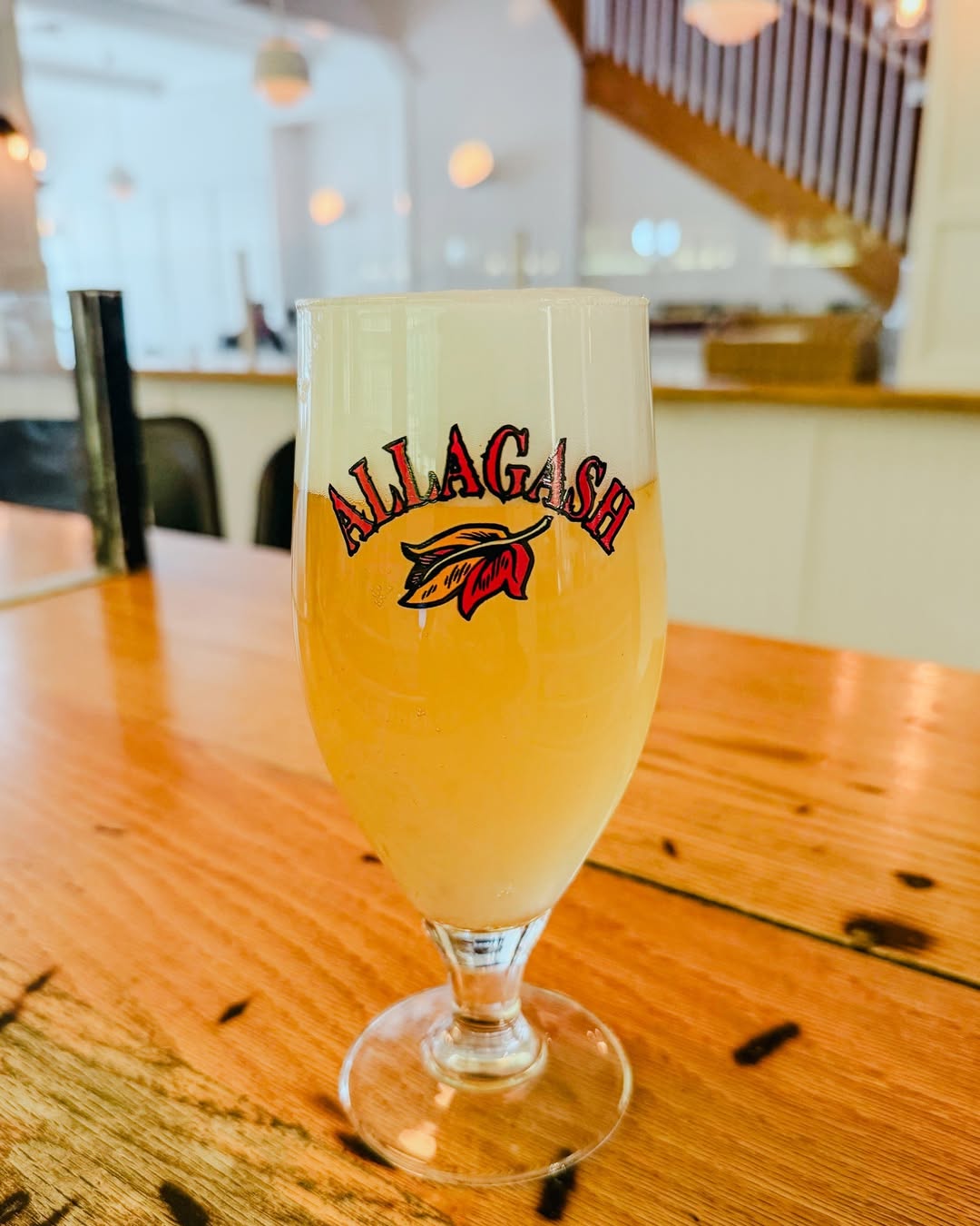 Glass of Allagash beer with foam on top, placed on a wooden table in a bright indoor setting.