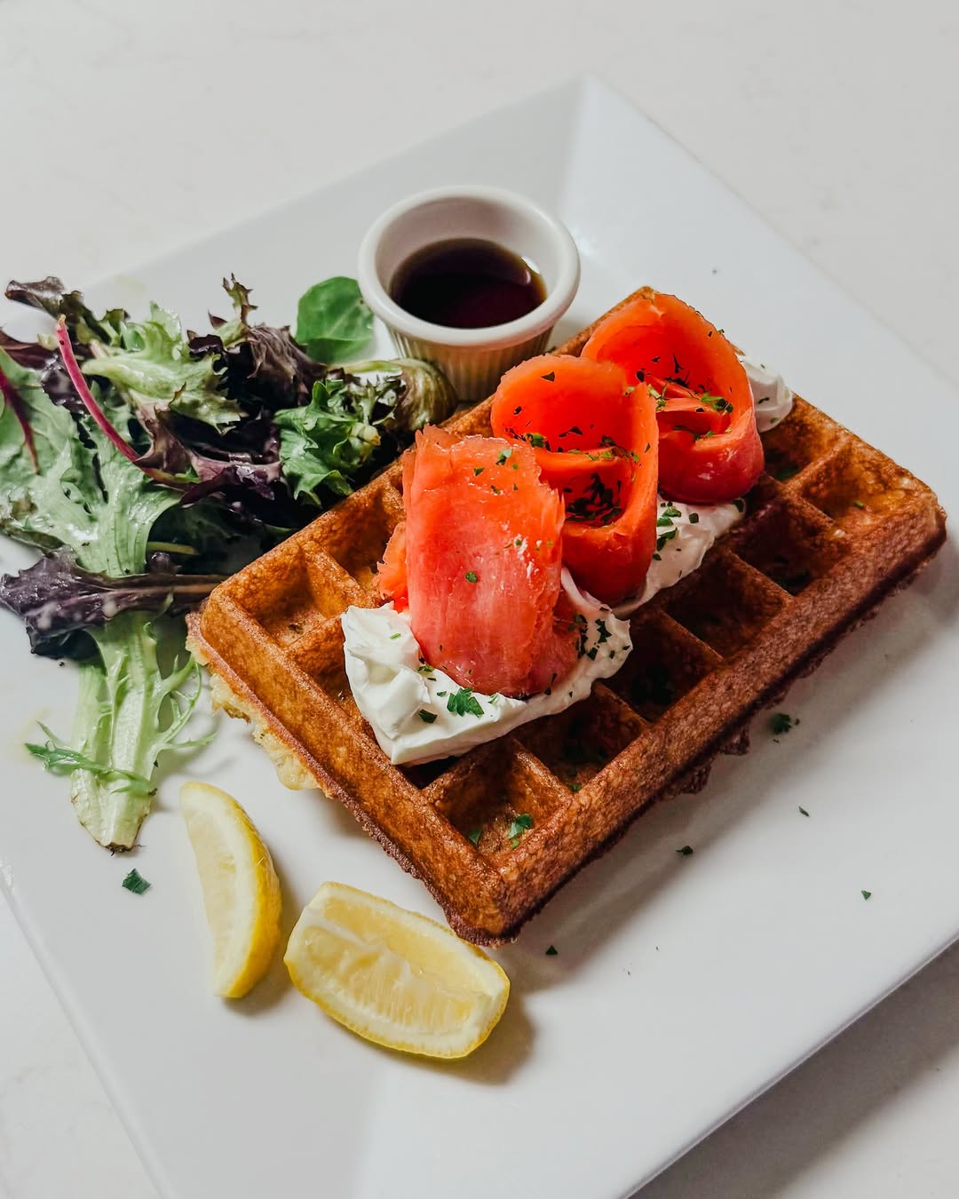Waffle topped with cream cheese and smoked salmon, served with mixed greens, lemon wedges, and a small cup of syrup on a white plate.