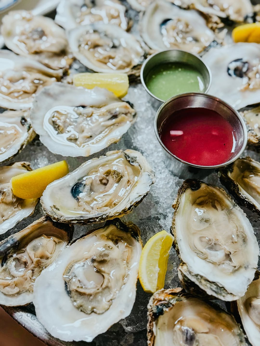 Tray of fresh oysters on ice with lemon wedges and two small metal cups of dipping sauces, one green and one red.