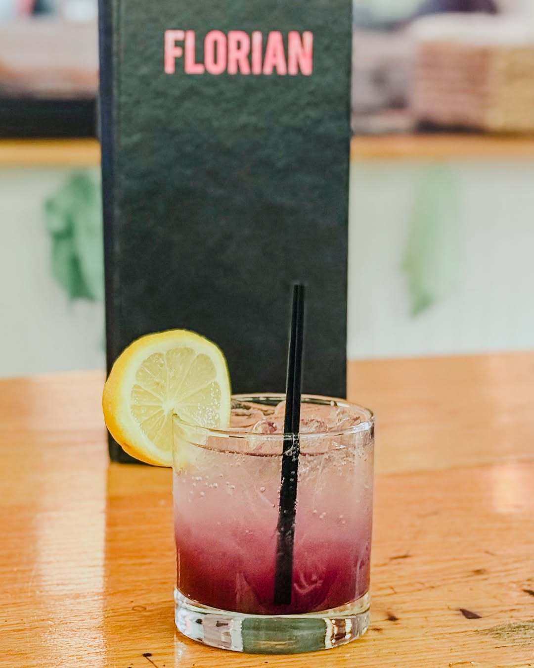 Glass of purple and clear iced drink with a lemon slice and black straw on a wooden table, with a black menu labeled FLORIAN in the background.